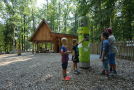 Kinder sehen sich eine Informationssäule vor einer Holzhütte im Wald an.