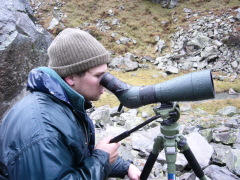 Ein Mann mit Mütze und blauer Regenjacke sieht durch ein Spektiv.